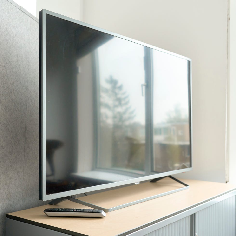 Best TV a large flat screen tv is turned off, but an image of a window is reflected, it is on a table where there is a control, in the background is a gray wall