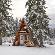 an attractive and stylish looking man stands and leans on the railing of the upstairs balcony of an a frame winter cabin the cabin is flanked by snow covered evergreens and a light snow fall blankets the scene a black rabbit can be seen outside the cabin