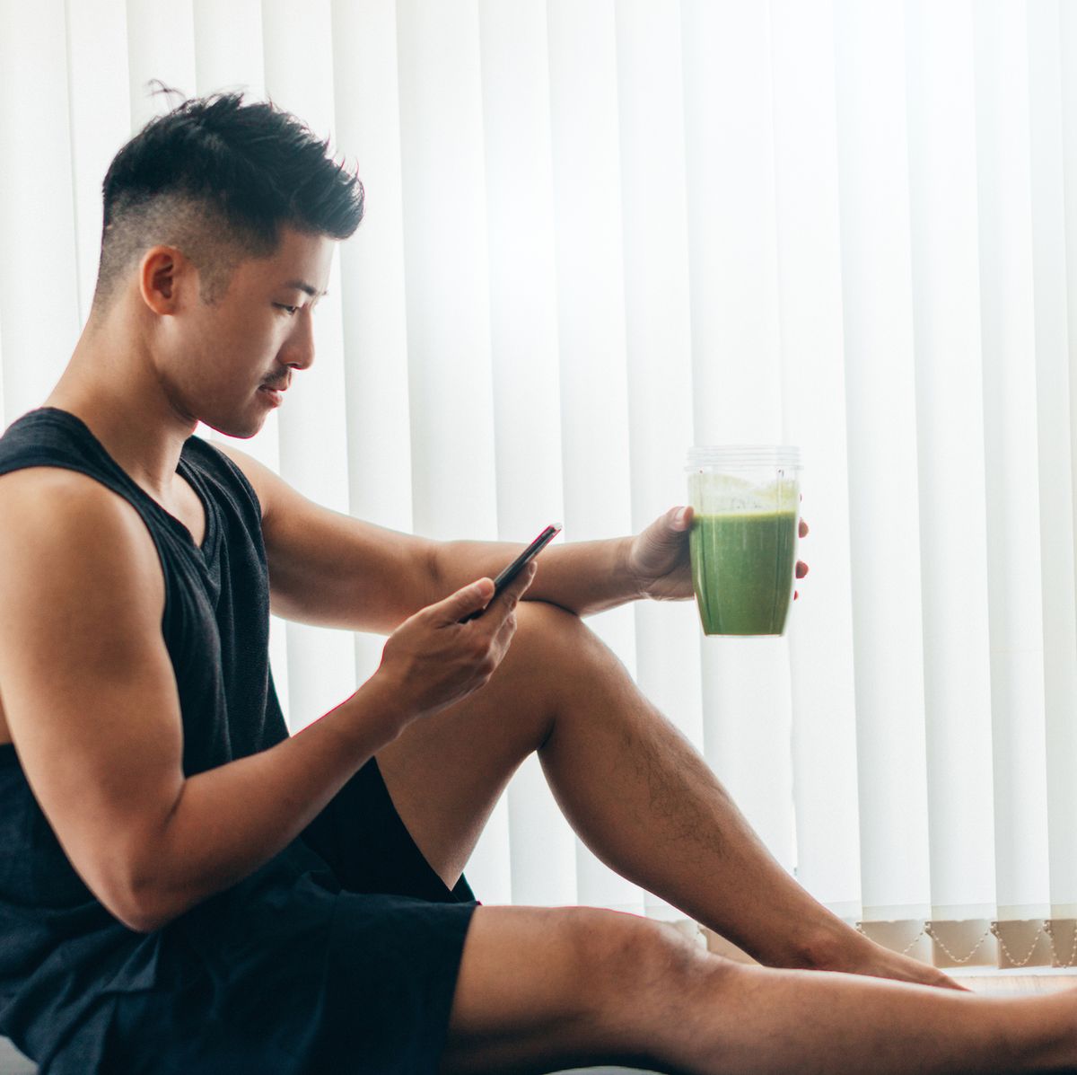 side view of a successful asian man in sportswear checking emails on smartphone while drinking green smoothie after exercise at home