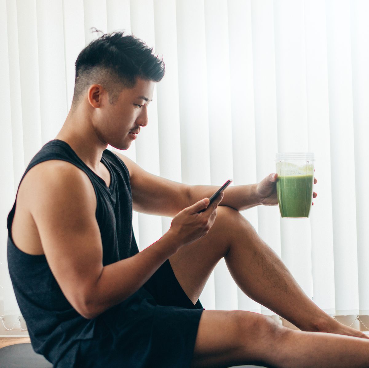 side view of a successful asian man in sportswear checking emails on smartphone while drinking green smoothie after exercise at home