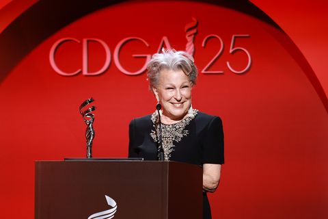 bette midler, wearing a black dress, stands at a podium with a microphone and a trophy, standing in front of a red wall