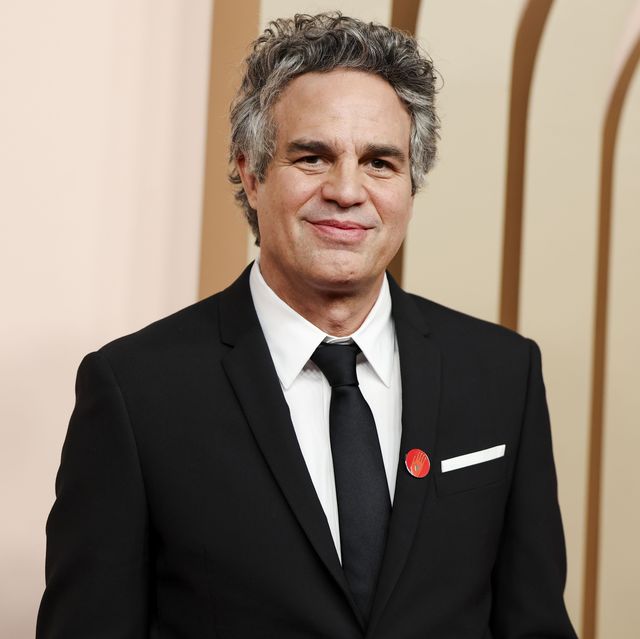 mark ruffalo smiles at the camera while standing in front of a tan background, he wears a black suit jacket and tie with a white collared shirt and a red button on his lapel