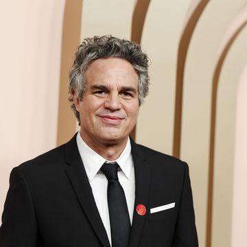 mark ruffalo smiles at the camera while standing in front of a tan background, he wears a black suit jacket and tie with a white collared shirt and a red button on his lapel