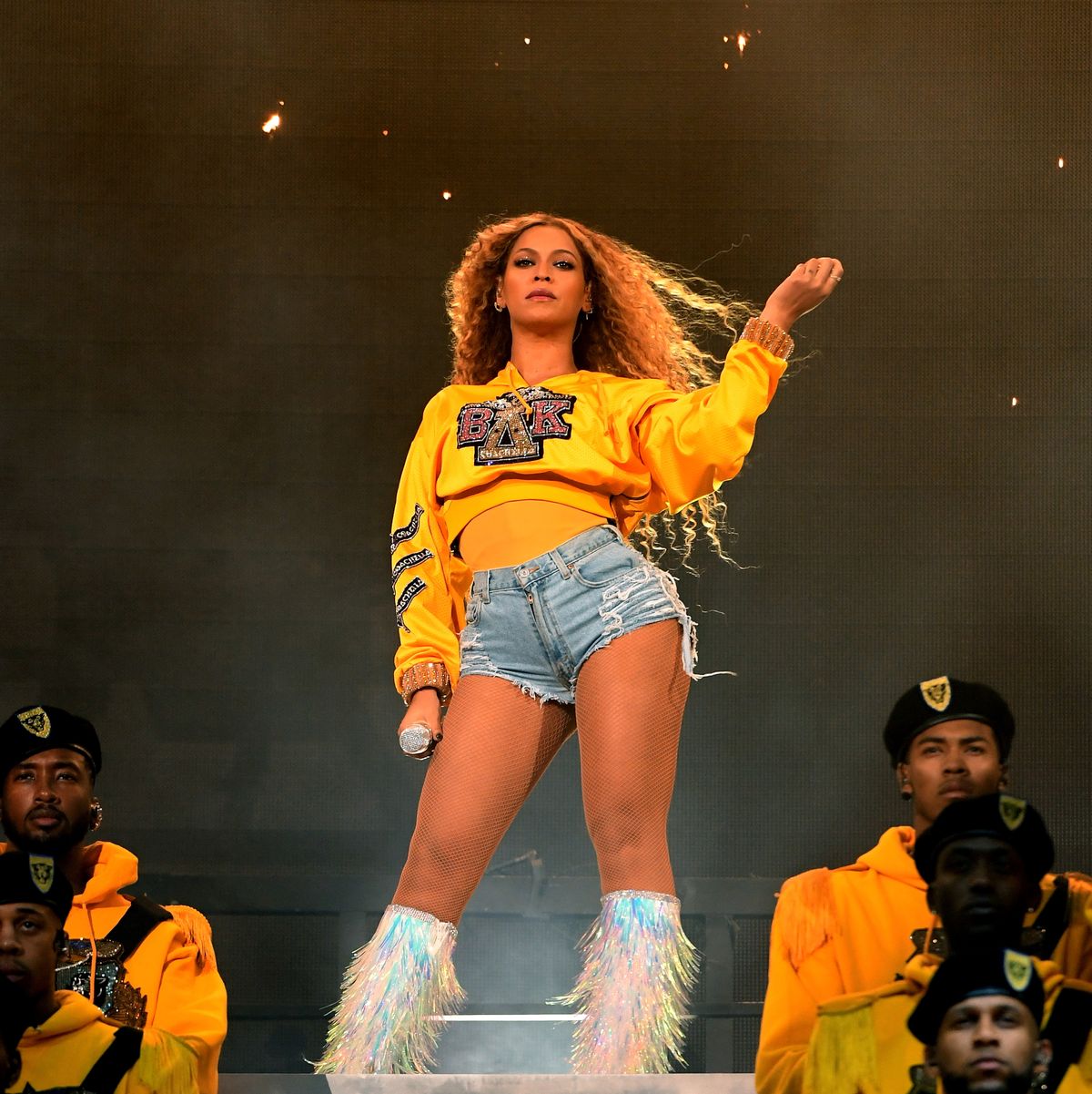beyonce stands on a stage with performers below her, she wears a yellow sweatshirt and jean shorts