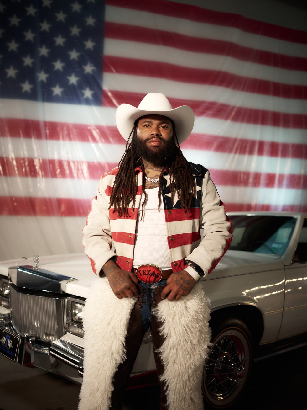 person in cowboy attire poses in front of a vintage car against an american flag backdrop