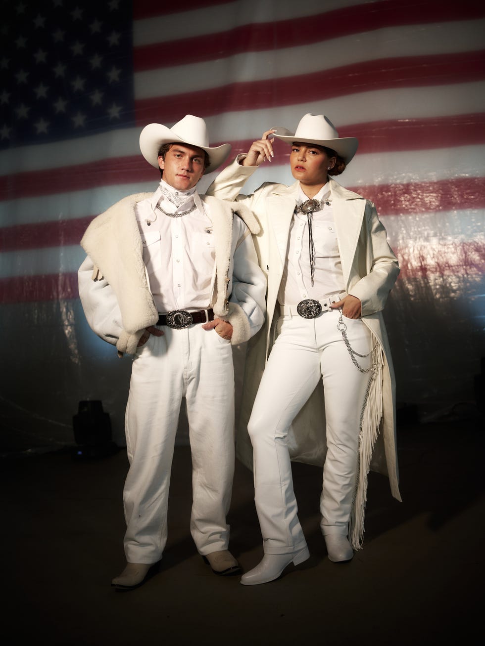 two individuals dressed in white cowboy attire in front of a flag backdrop
