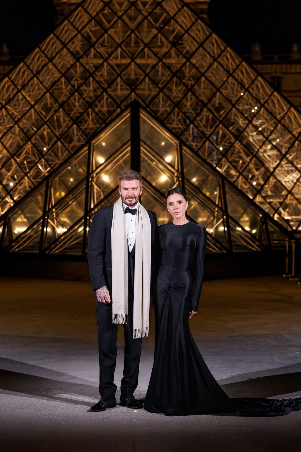 david and victoria beckham posing elegantly in front of a glass pyramid structure at night