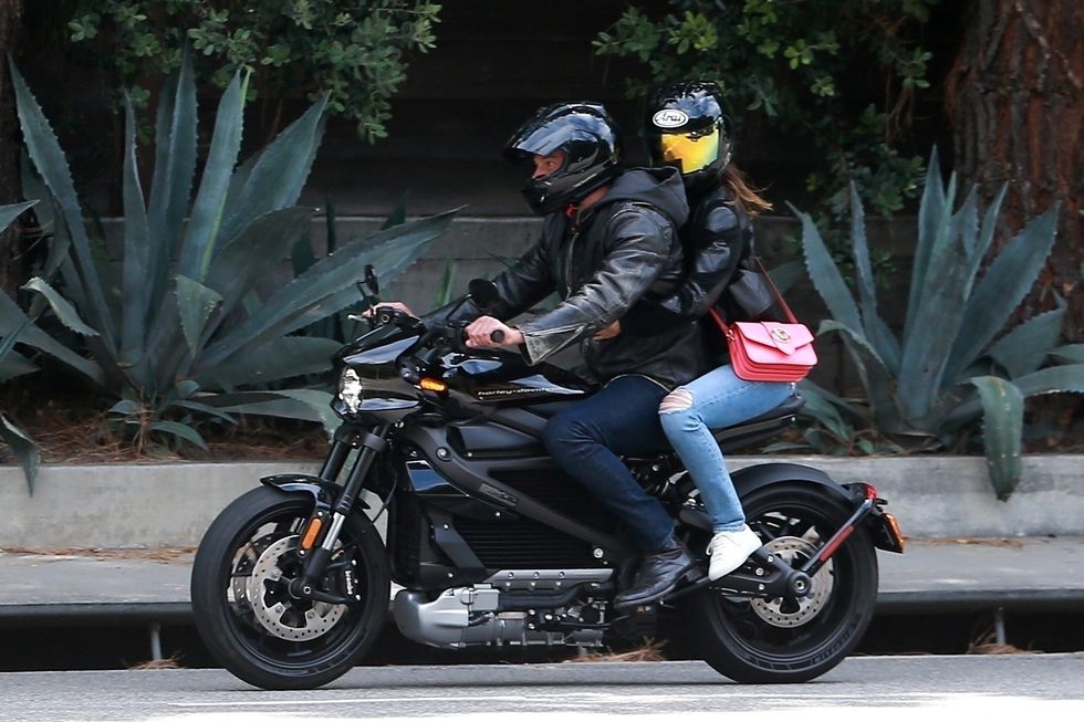 the couple sits on a black motorcycle, both wearing black helmets and black leather jackets