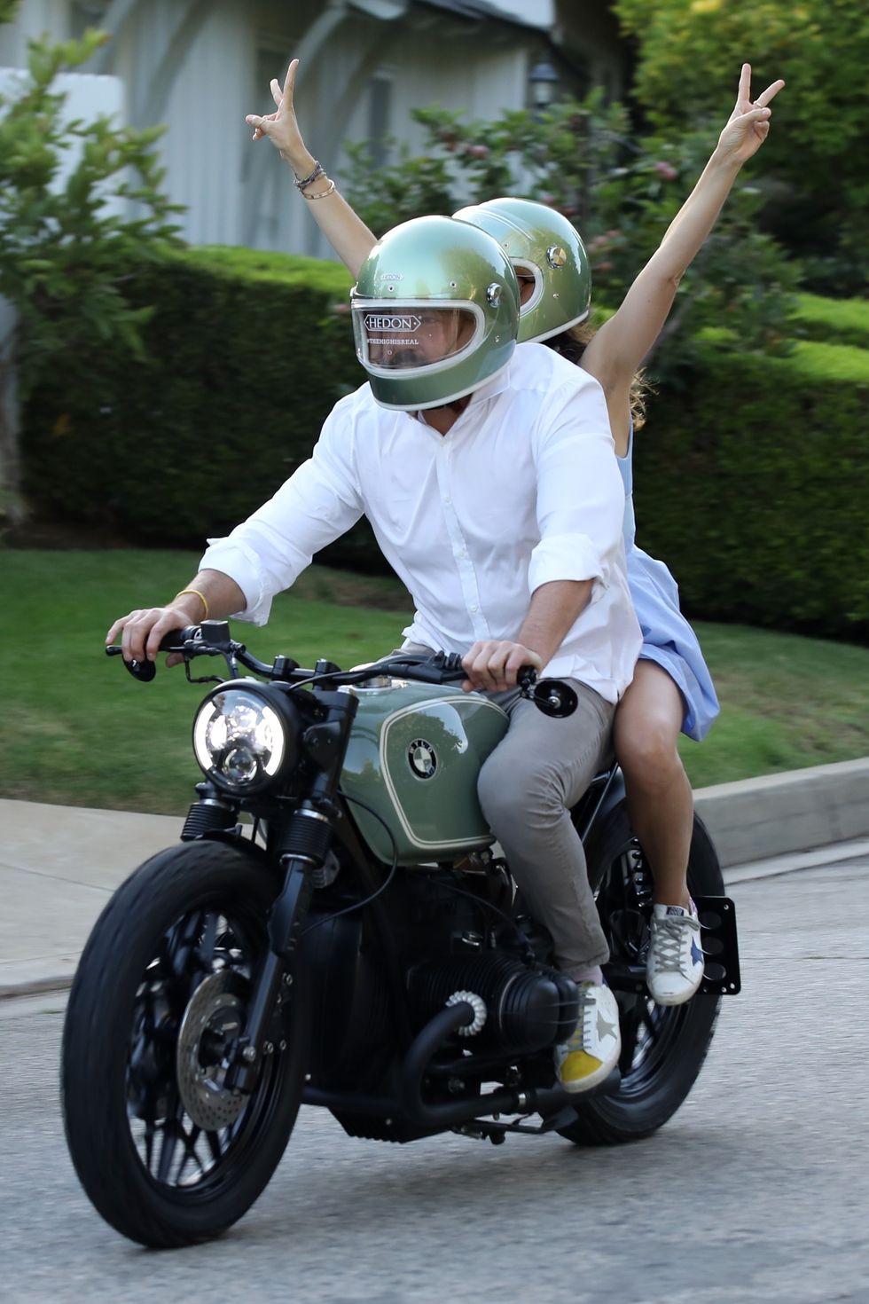 ben and ana wearing green motorcycle helmets while riding a green bmw motorcycle through a neighborhood ana throws both her hands up in the back to gesture peace signs
