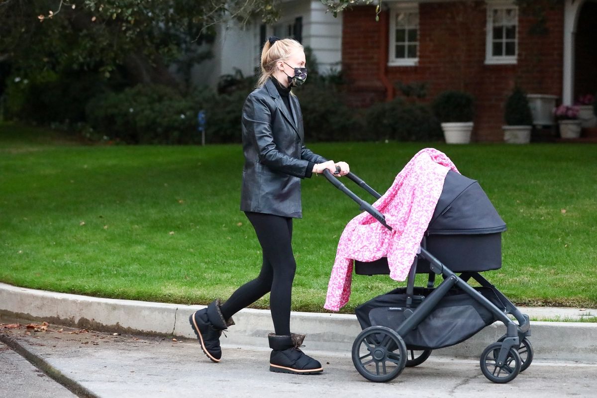 sophie turner rounds a corner on a neighborhood dressed in all black while pushing a black baby stroller
