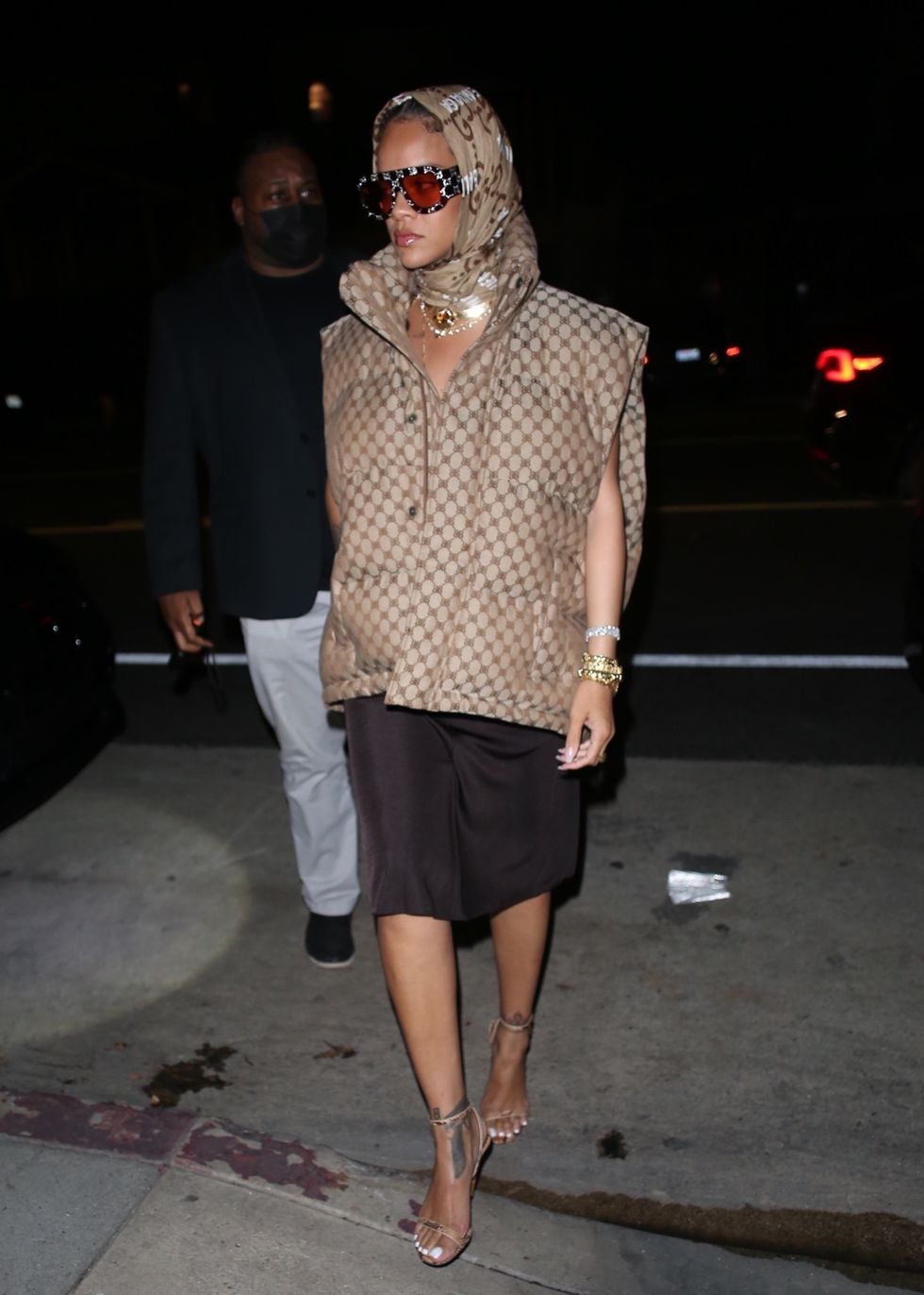 rihanna walks at night wearing padded vest and monogrammed head scarf