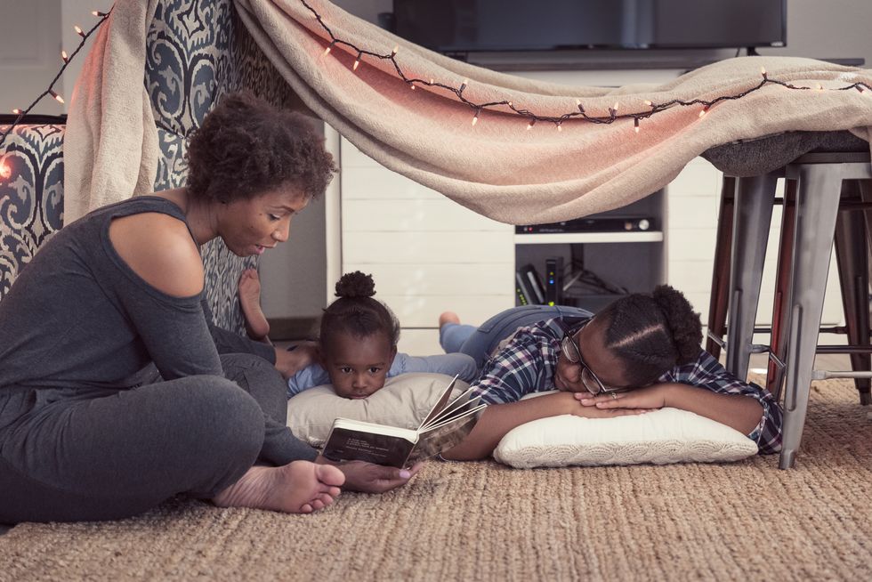 mom sitting until a fort made of blankets with her two daughters, reading a story from a book