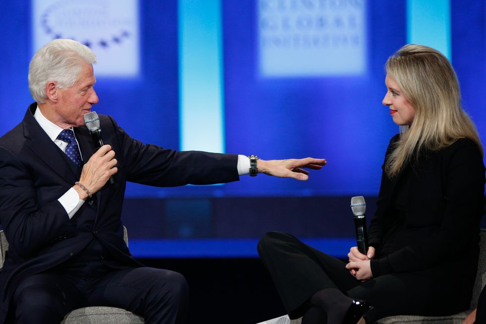 bill clinton speaks into a microphone while looking at and extending his arm out to elizabeth holmes who is looking at him and holding a microphone in her hands, both are sitting on a stage with a blue background behind them