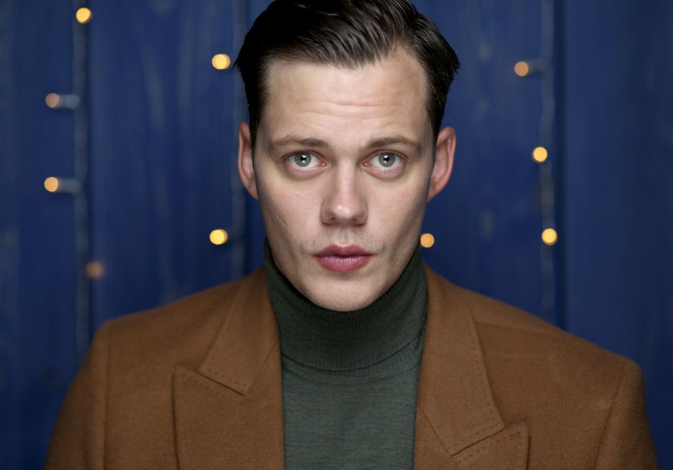 bill skarsgard stands in front of a bright blue wall with fairy lights hanging behind him