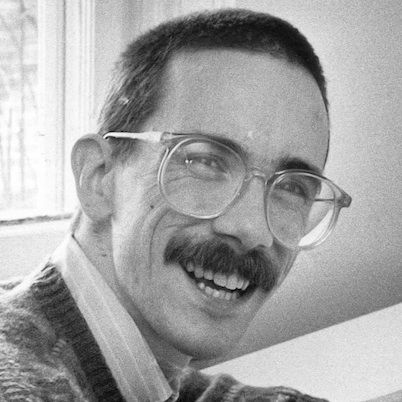 bill watterson smiles at the camera while looking over one shoulder, he wears large circular glasses a collared shirt and a sweater