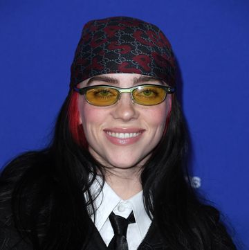 billie eilish smiles at the camera, she wears a black jacket over a white collared shirt and black tie with yellow tinted glasses and a patterned black and red bandana