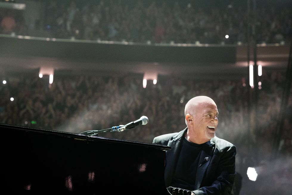 a man sits at a piano outfitted with a microphone and looks to the right as he smiles, a blurred crowd cheers in the background