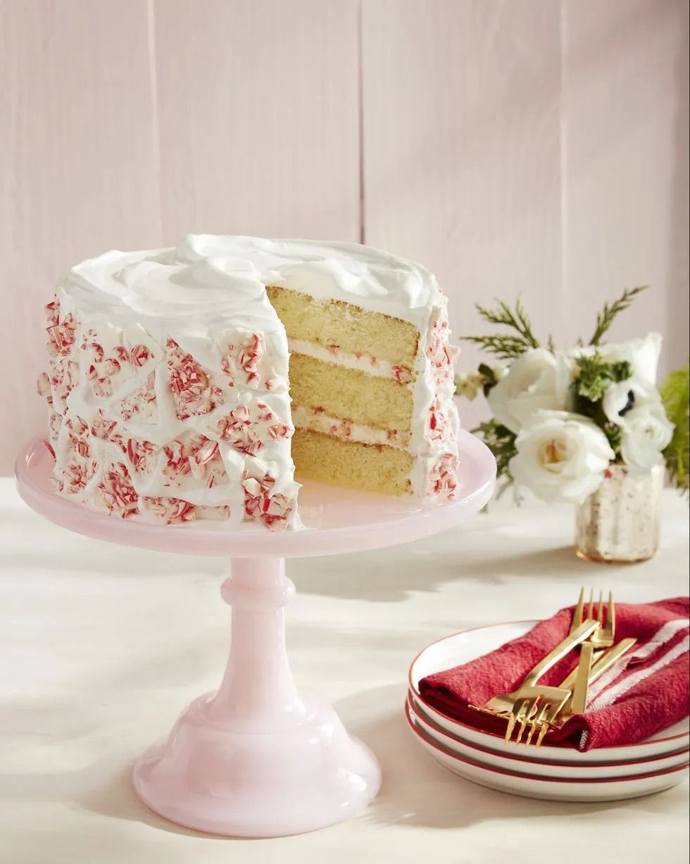 candy cane layer cake with white frosting and peppermint bark around the outside on a light pink cake stand