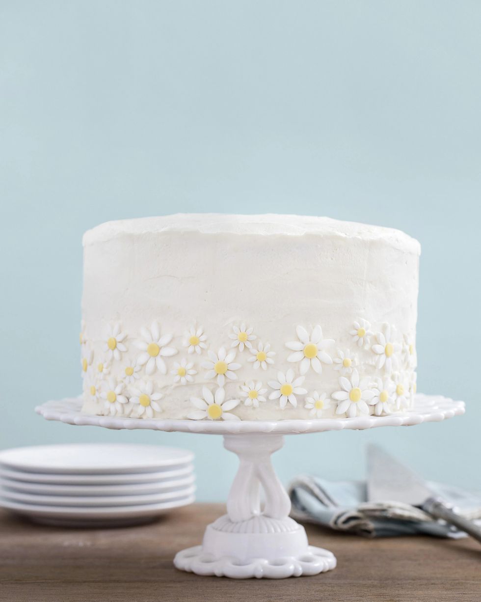 lemon coconut cake covered in white frosting with sugar daisies around the outside on a white cake stand