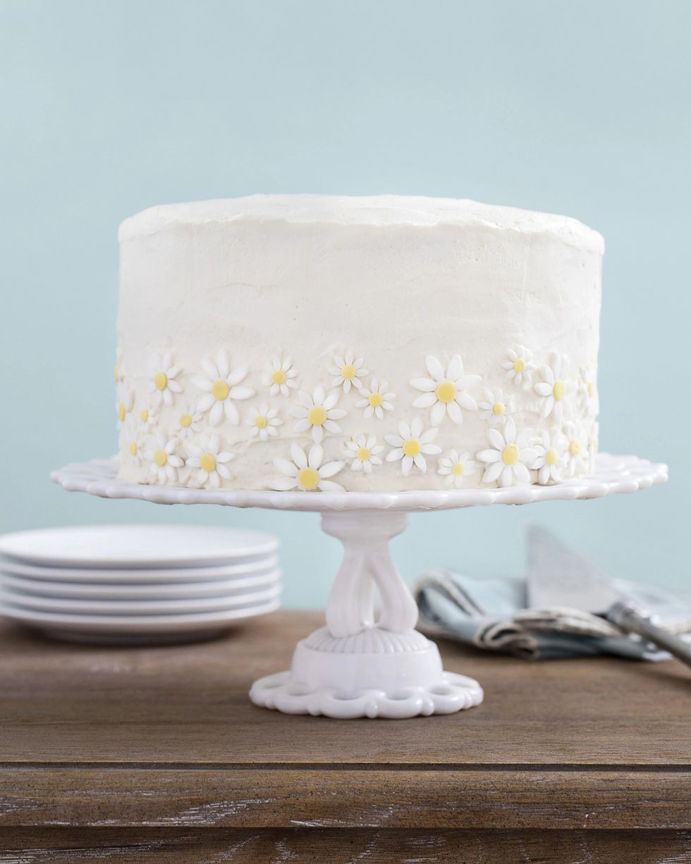 lemon coconut cake covered in white frosting with sugar daisies around the outside on a white cake stand