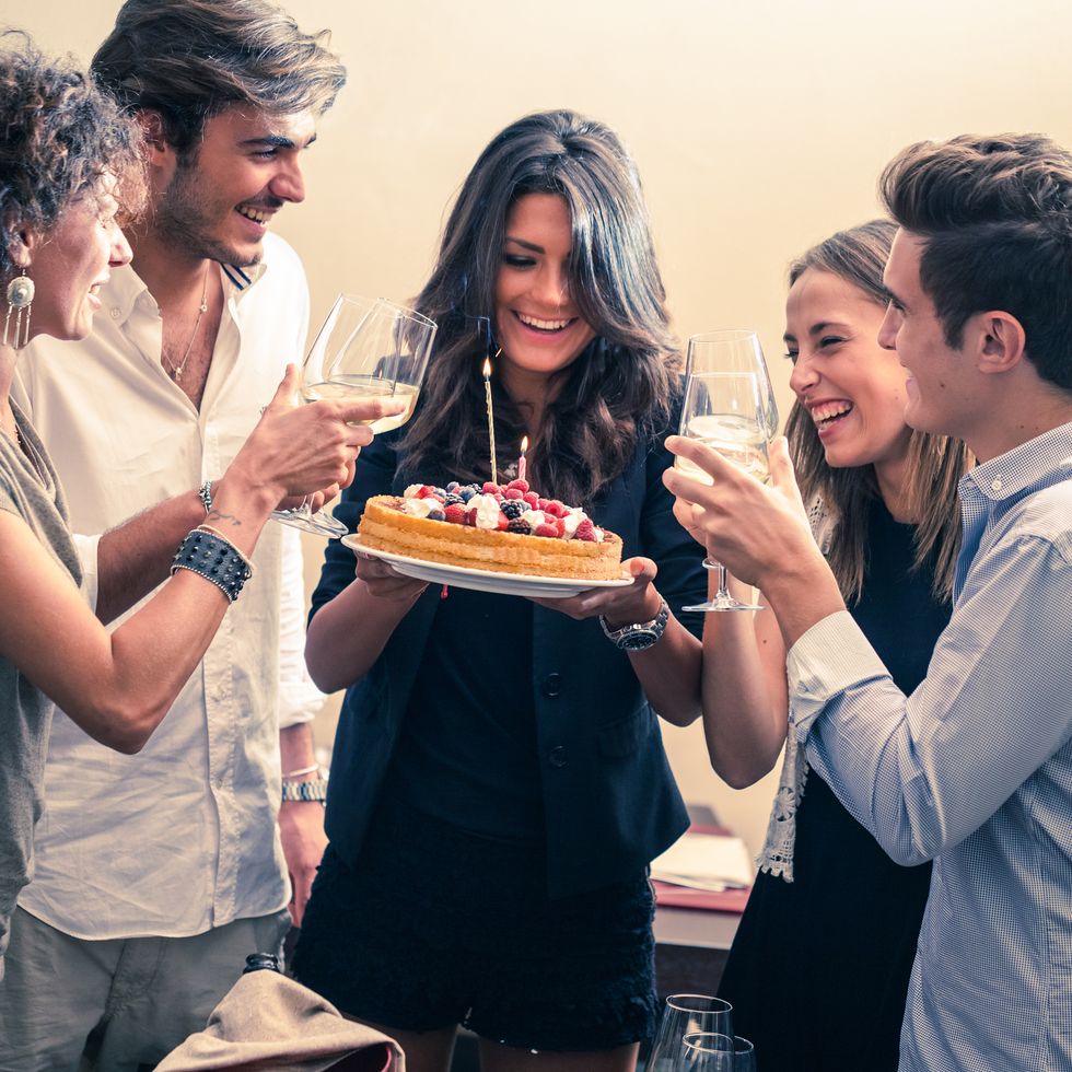a white woman in black holding a cake with fruit on top and a candle surrounded by friends holding wine glasses