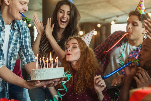 birthday girl blowing candles on a birthday cake and making wish surrounded by friends with party hats blowing streamers