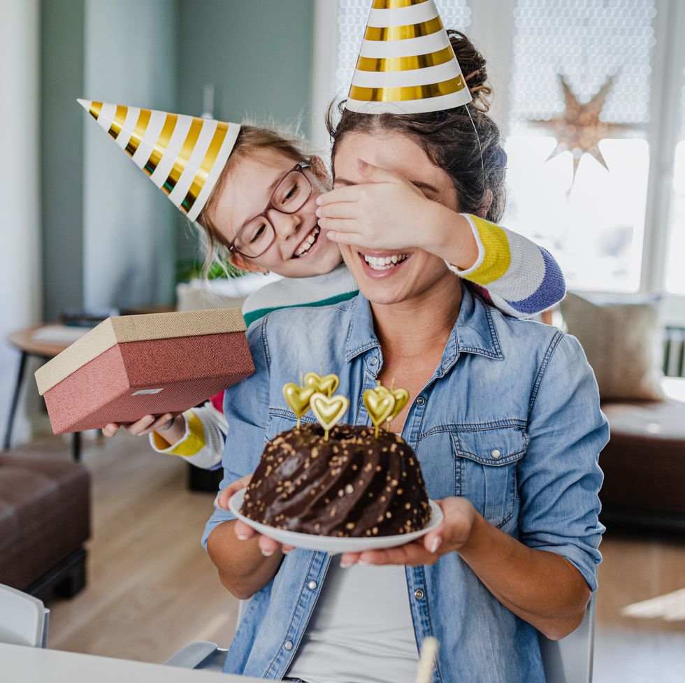 young mother and daughter are at home, wearing party hats and holding a birthday cake as they celebrate mothers birthday