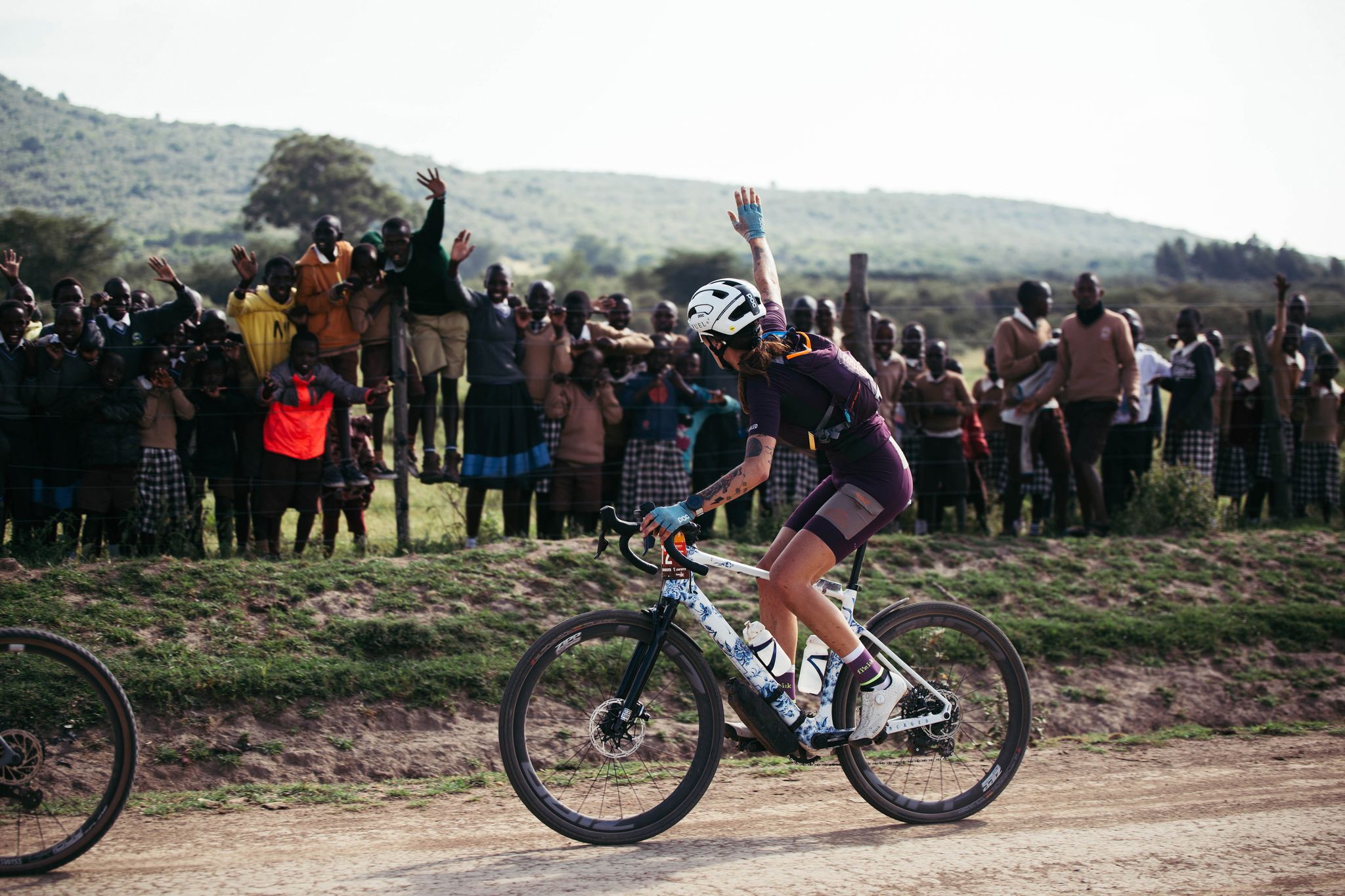 a cyclist riding on a dirt road waves to a crowd of spectators