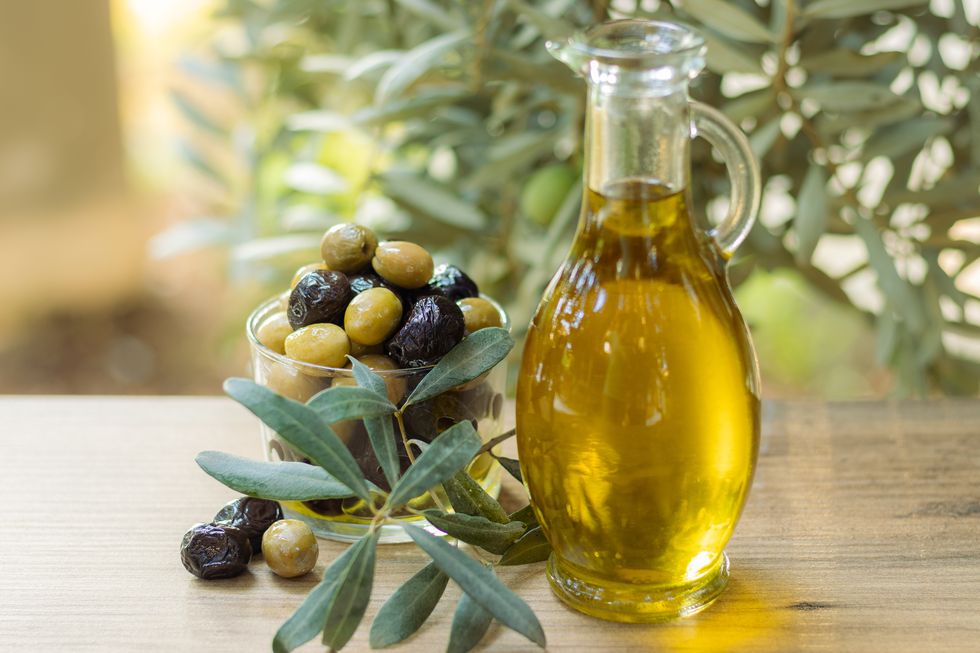 black and green olives in a transparent bowl next to an olive oil in a bottle in front of an olive tree