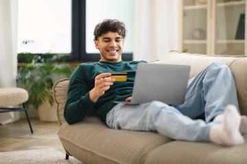 a young man smiles as he sits on a sofa and uses a card to pay for shopping on a laptop