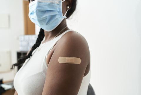 black woman with braid in her hair with white shirt and mask showing the band aid on her arm after vaccination, with white background concept of covid