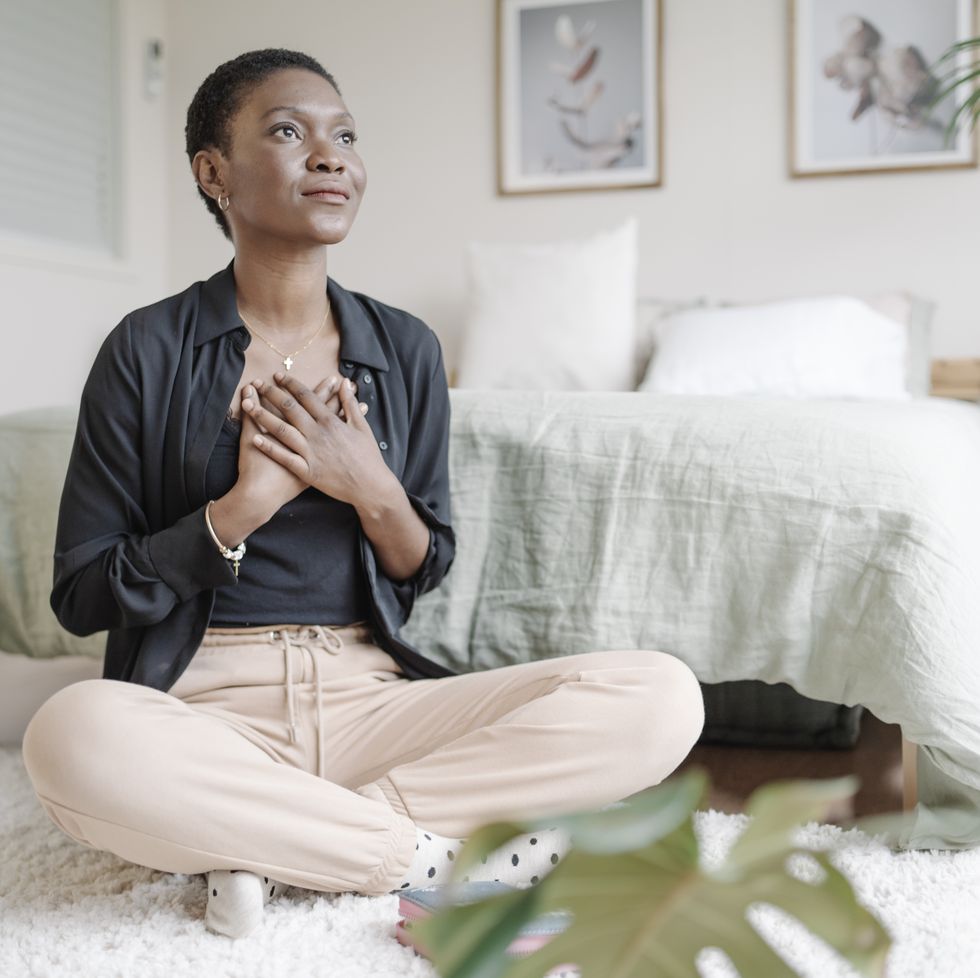 black woman with hand on chest showing gratitude while praying sitting on the floor in her bedroom.