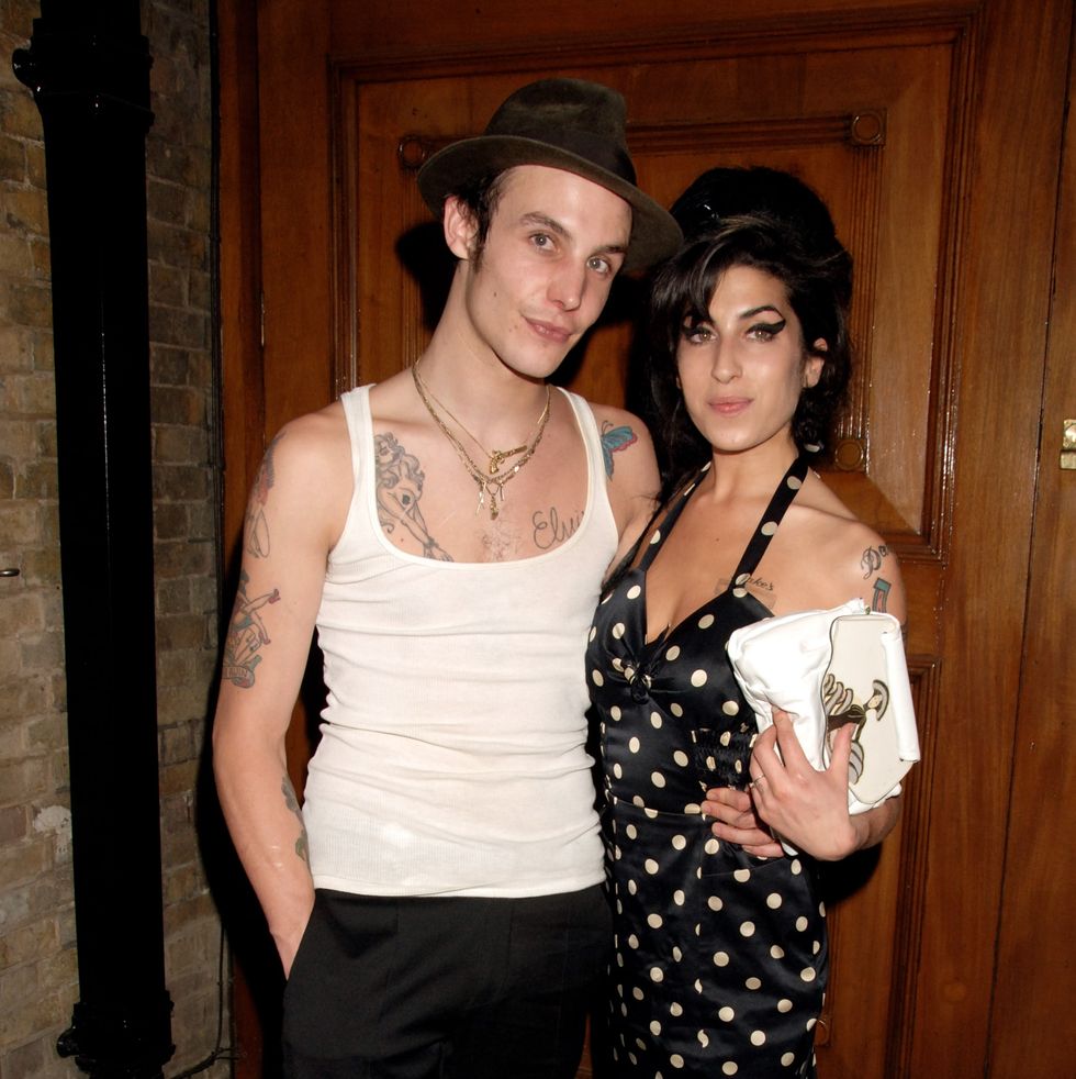 blake fielder civil and amy winehouse embrace while standing and posing for a photo, both smile softly as they stand in front of a wooden paneled wall, he wears a white tank top and black pants with a dark hat and gold necklaces, she wears a black halter dress with white polka dots and holds a white clutch in one hand
