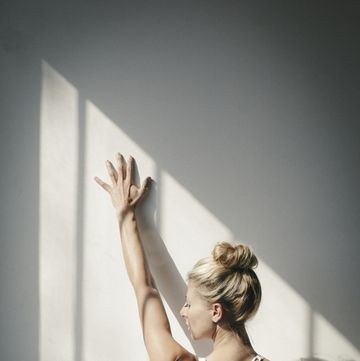 a blonde woman, in a white crop top and leggings, standing in front of a white wall, doing yoga, her arm raised, touching the wall
