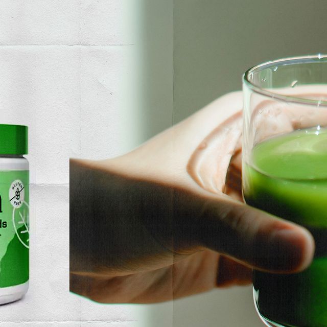 a hand holding a glass of green liquid next to a tub of bloom greens and superfoods