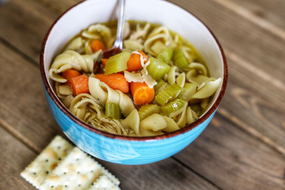 a blue bowl of homemade organic chicken noodle soup with vegetables and crackers on a wooden table