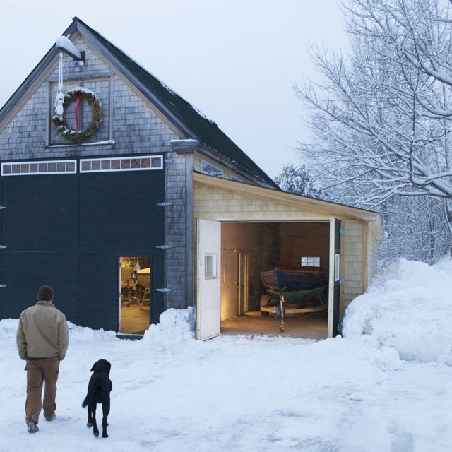A boat builder,  with his dog at his side, walk through the snowy yard after a long winters day in the boat shed.