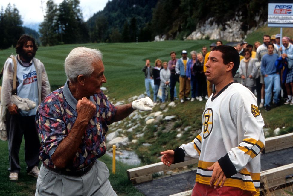 bob barker and adam sandler stand and face each other, barker wears a patterned collared shirt and gray khakis and has his fists raised, sandler wears a white boston bruins jersey and appears to back away from barker, a crowd of people standing on a golf course are in the background
