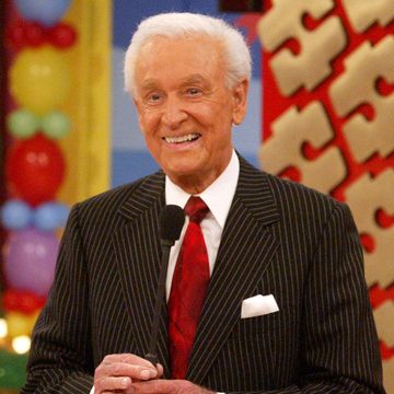 bob barker smiles while holding a microphone, he stands in front of a tv set with multicolored balloons and wears a black pinstriped suit jacket with a white collared shirt, red tie, and white pocket square