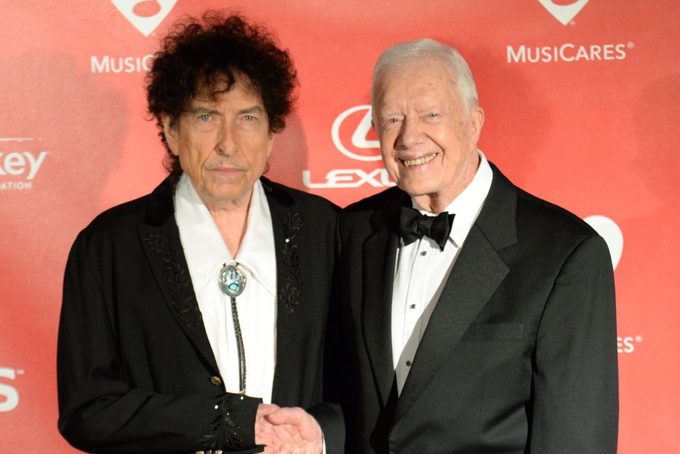 bob dylan, unsmiling, wearing a black suit jacket and white shirt, shaking hands with jimmy carter, smiling, wearing a black tuxedo
