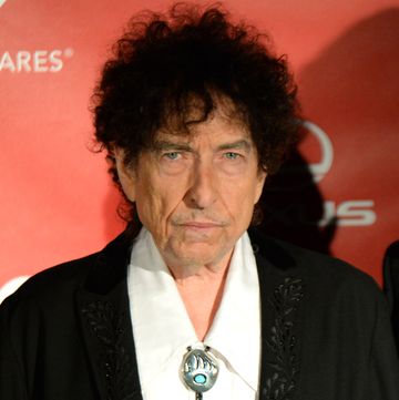 bob dylan looks at the camera, he wears a black suit jacket and white collared shirt with a silver bolo tie