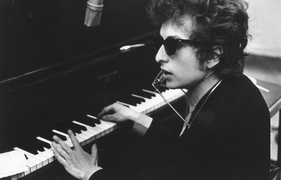 Bob Dylan sits at the piano, wearing sunglasses and a harmonica around his neck