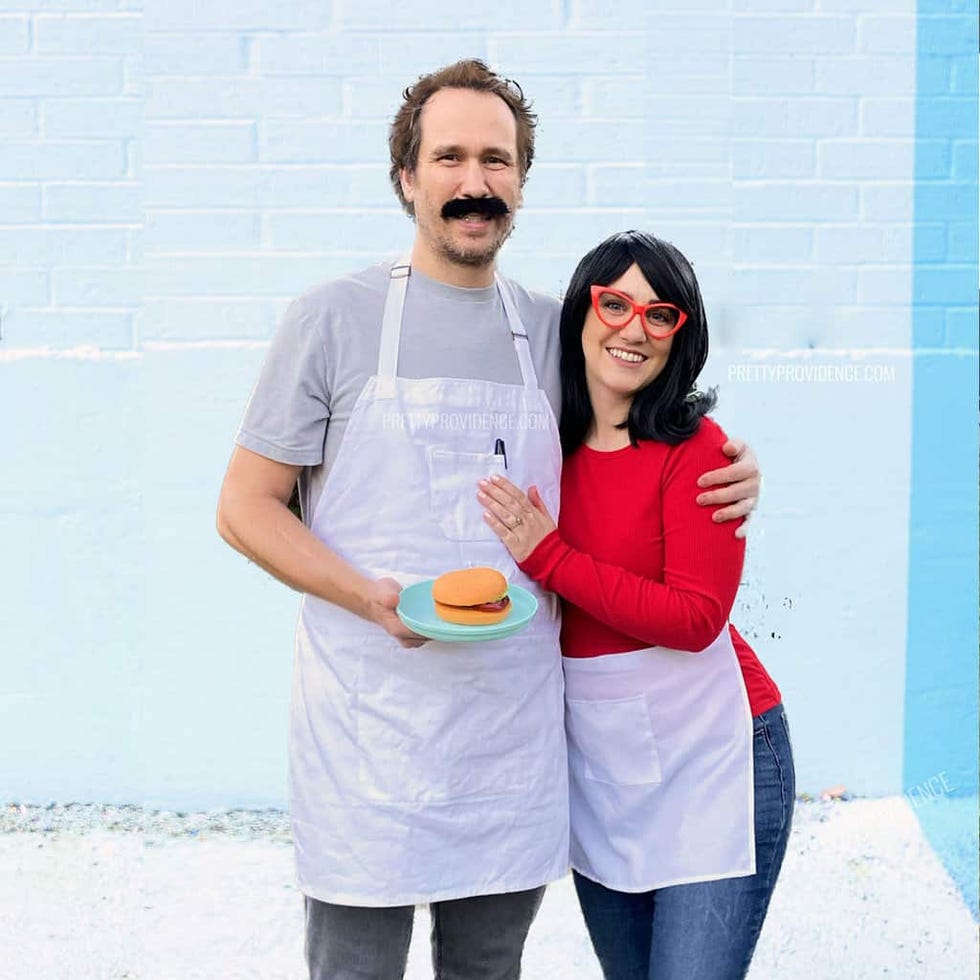 two individuals in aprons holding a burger on a plate against a blue wall