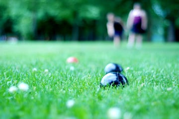 two black bocce balls on green lawn with red ball behind them out of focus and two players further back also out of focus