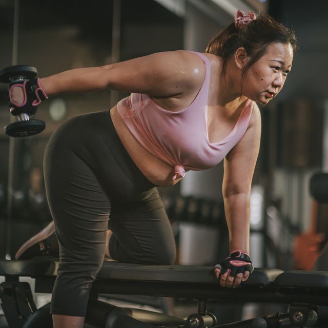 body positive asian mid adult woman exercising with dumbbells in a lunge position at gym bench at night