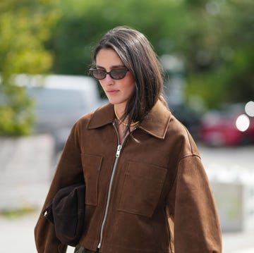 bolso mujer una mujer lleva el cabello liso castaño oscuro hasta los hombros, con la raya ligeramente descentrada, gafas de sol rectangulares negras estrechas con lentes oscuras, un collar corto de cuentas amarillas superpuesto con un collar de cadena fina de plata, una chaqueta corta de ante marrón chocolate con cuello abierto, cremallera plateada expuesta en la parte delantera, dos grandes bolsillos de parche en el pecho, hombros caídos y mangas anchas, un bolso de mano blando oscuro doblado y llevado bajo el brazo, y unos pantalones verde oliva apenas visibles.