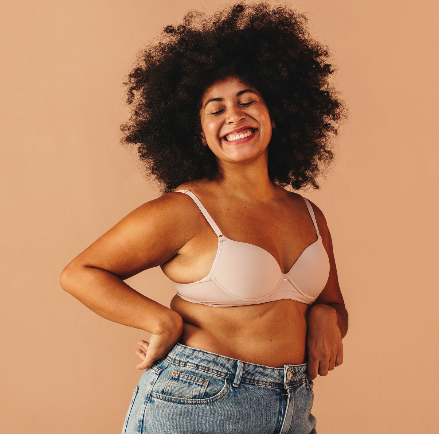 self confident young woman smiling happily while wearing a bra and jeans in a studio