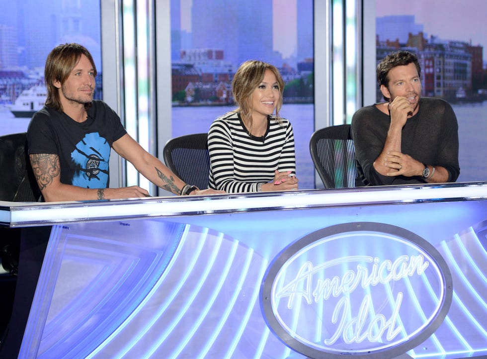 keith urban, jennifer lopez, and harry connick jr sit behind a table with the american idol logo on it and listen as someone off camera performs