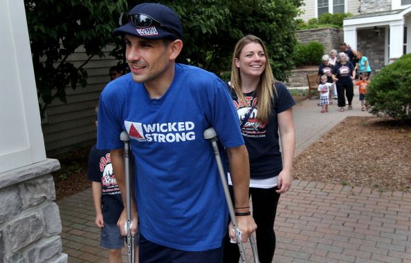 marc fucarile wearing a blue t shirt with the words wicked strong on it, using crutches and smiling, walking in front of a house with a smiling woman walking behind him