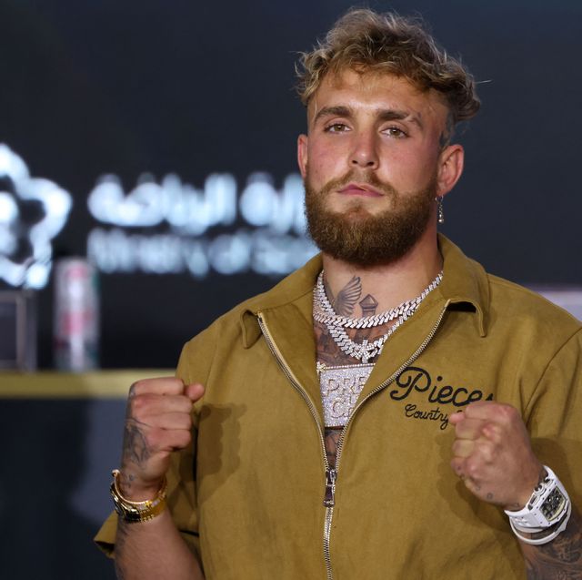 jake paul poses with two fists held at his chest, he wears a mustard yellow jacket and big necklaces